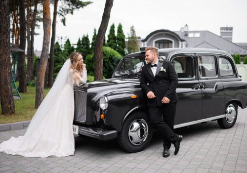 Side view of stylish and handsome groom in black tuxedo, leaning to retro car and looking to bride, which standing opposite and lovely looking to him during wedding walk on open air